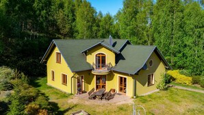 Una casa de dos pisos con balcón, rodeada de un césped cuidado y un bosque frondoso.