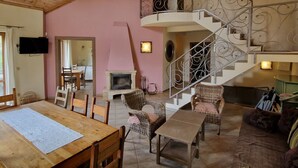 Un amplio salón con techo de madera, chimenea, muebles de mimbre, una escalera con barandilla decorada y un televisor montado en la pared.