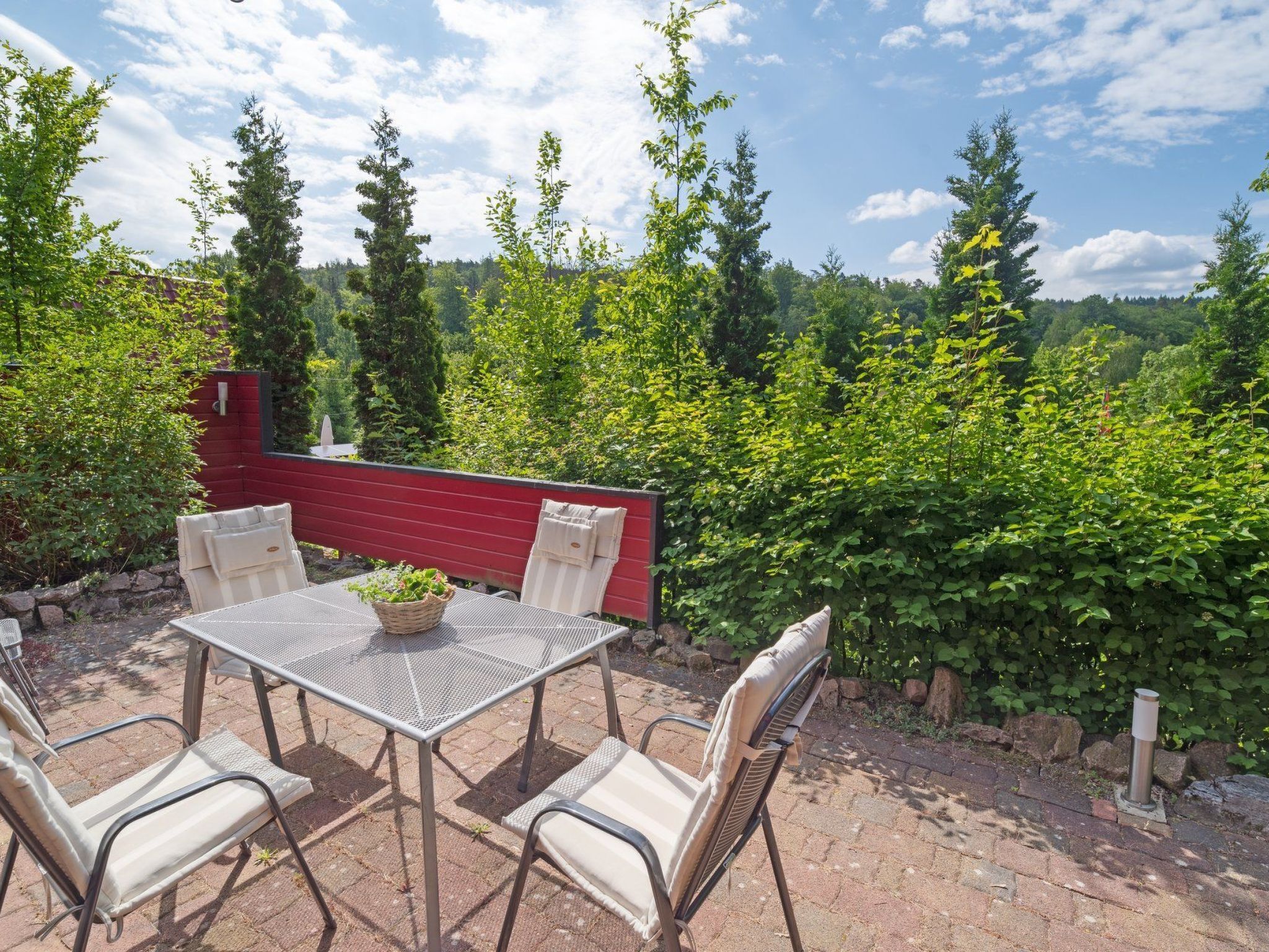 Une terrasse extérieure avec une table et des chaises, entourée de verdure et bordée d’un mur rouge.