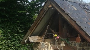 Una rustica casa in pietra con tetto spiovente, una porta in legno e una finestra. Sono presenti aree salotto all'aperto con sedie e un tavolo, immerse nel ghiaione e tra le piante.