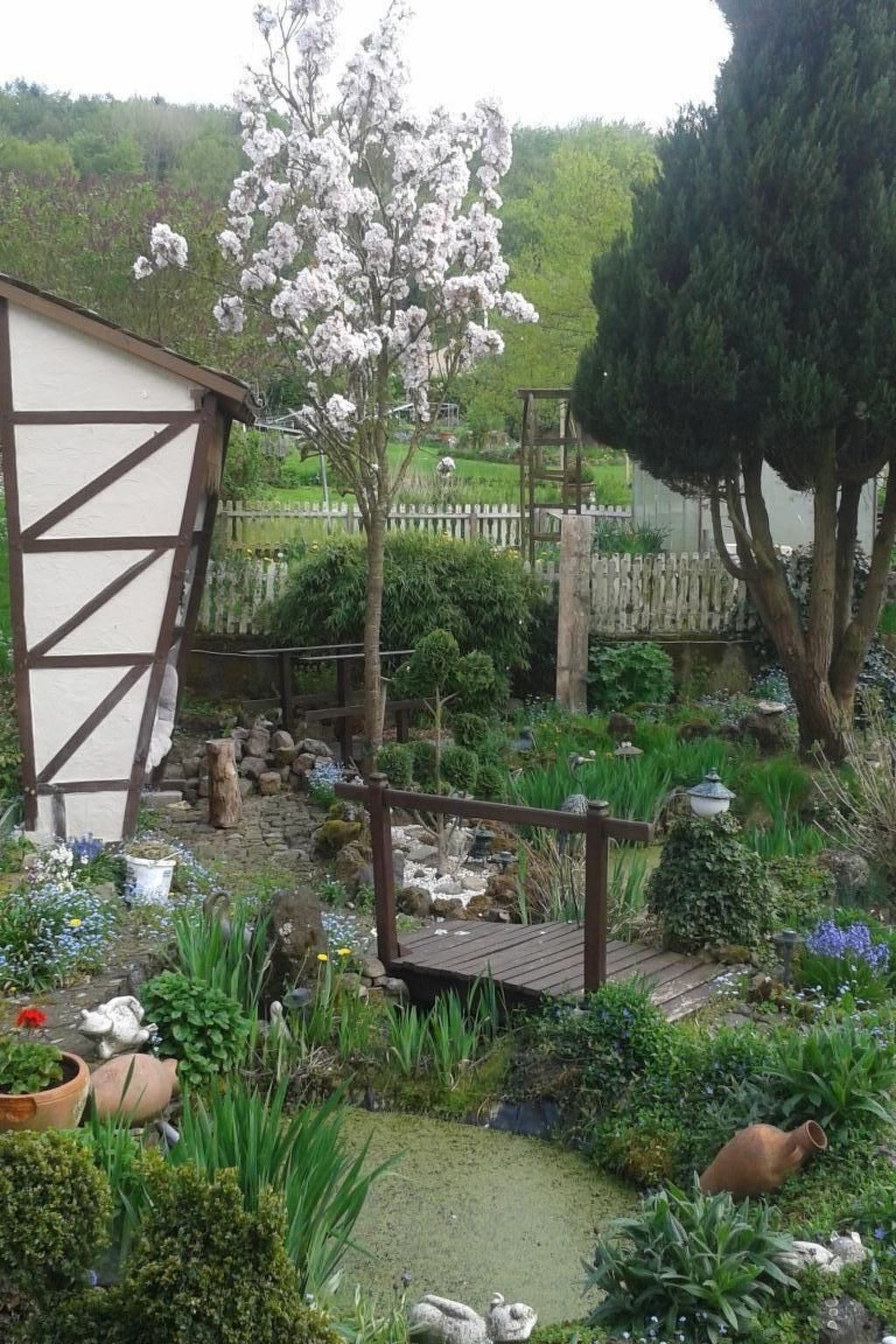 Un jardin agrémenté d’une terrasse en bois, un bâtiment blanc à la structure en bois apparent, et diverses plantes et fleurs.