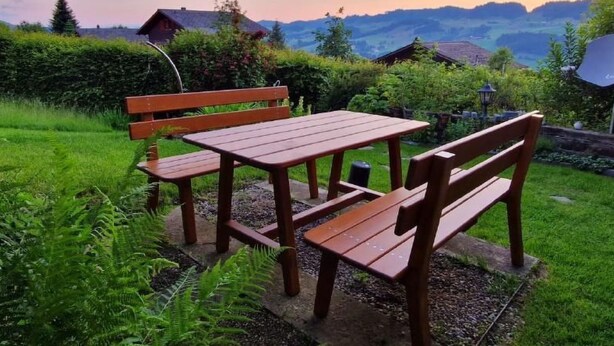 Un banc et une table en bois, installés dans un jardin luxuriant, offrent une vue sur les montagnes et une maison qui se dessine à l’arrière-plan.