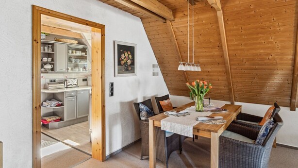 Zona pranzo con tavolo in legno, sedie in vimini e un vaso di fiori. Si vede una cucina attraverso un'apertura.