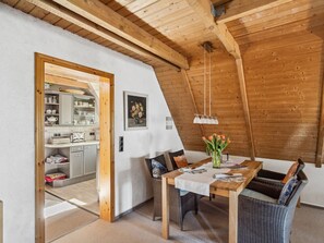 A dining area with a wooden table, wicker chairs, and a vase of flowers. There is a kitchen visible through an open doorway. - Pellworm by Interhome (Pellworm (Ort))