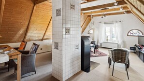 Un'ampia stanza mansardata con travi in legno, un termosifone nero, un tavolo da pranzo con sedie e un'ampia finestra con tende.