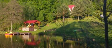 Una cabaña junto al lago con techo rojo, rodeada de un bosque frondoso y un cielo despejado.