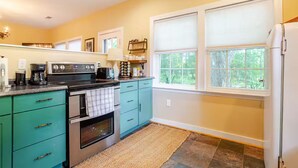 Una cocina con armarios azul verdoso, una estufa y un refrigerador. Hay ventanas con persianas, una alfombra en el suelo y un estante con utensilios de cocina.