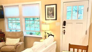 Una sala de estar acogedora con un sofá, una mesa de comedor y una silla. Hay ventanas con persianas, una puerta y un cuadro enmarcado en la pared.