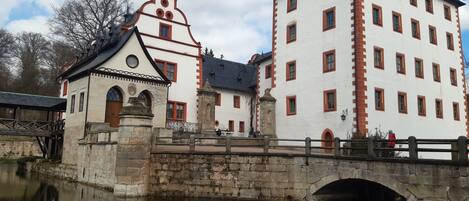 A historic building with a white facade and red accents, featuring a prominent clock tower and a bridge over a calm water body.