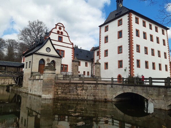 A historic building with a white facade and red accents, featuring a prominent clock tower and a bridge over a calm water body. - zum Spaal by Interhome (Uhlstädt-Kirchhasel)
