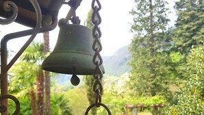A bell hanging outdoors, possibly part of a garden or park setting with greenery and trees in the background. - Casa Elena by Interhome (Locarno Tegna)