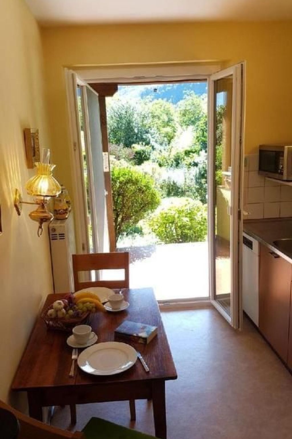 A dining area with a table set for two, a lamp, and a view of a garden through an open door.