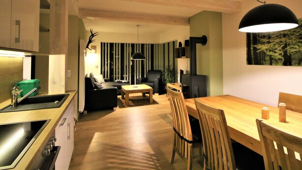 A modern kitchen with a built-in stove, a dining area with a wooden table and chairs, a black sofa, and a large window with vertical blinds. - Moderne, niveauvoll eingerichtete Ferienwohnung by Interhome (Carlsfeld)