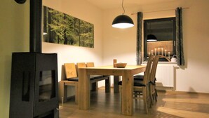 A dining area with a wooden table and chairs, a black stove, a hanging light, and a window with curtains. - Moderne, niveauvoll eingerichtete Ferienwohnung by Interhome (Carlsfeld)