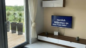 Ein modernes Wohnzimmer mit großem Fernseher, der eine Willkommensnachricht zeigt, einem hölzernen Konsolentisch und Blick auf topfpflanzte Outdoor-Bepflanzung durch eine Glastür.