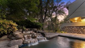 A serene outdoor setting with a small waterfall, stone steps, and a pool, surrounded by trees and a stone wall. - Montgomery Ranch | Stunning Lake House (Redding)