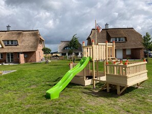 A playground with a green slide and wooden play structures, set against houses with thatched roofs. - Vacation home Knut by Interhome (Groß Schoritz)