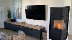 Uma sala de estar moderna com uma televisão de tela plana na parede, uma mesa de consola de madeira com uma planta, um quadro e uma lareira a lenha preta.