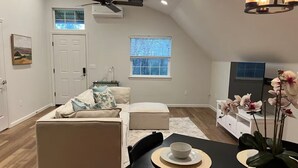 A modern loft-style apartment with a dining area, a living room with a sofa and ottoman, a kitchen area, and a large window providing natural light. - Peaceful Lakeside Apartment Garage & EV (Redding)