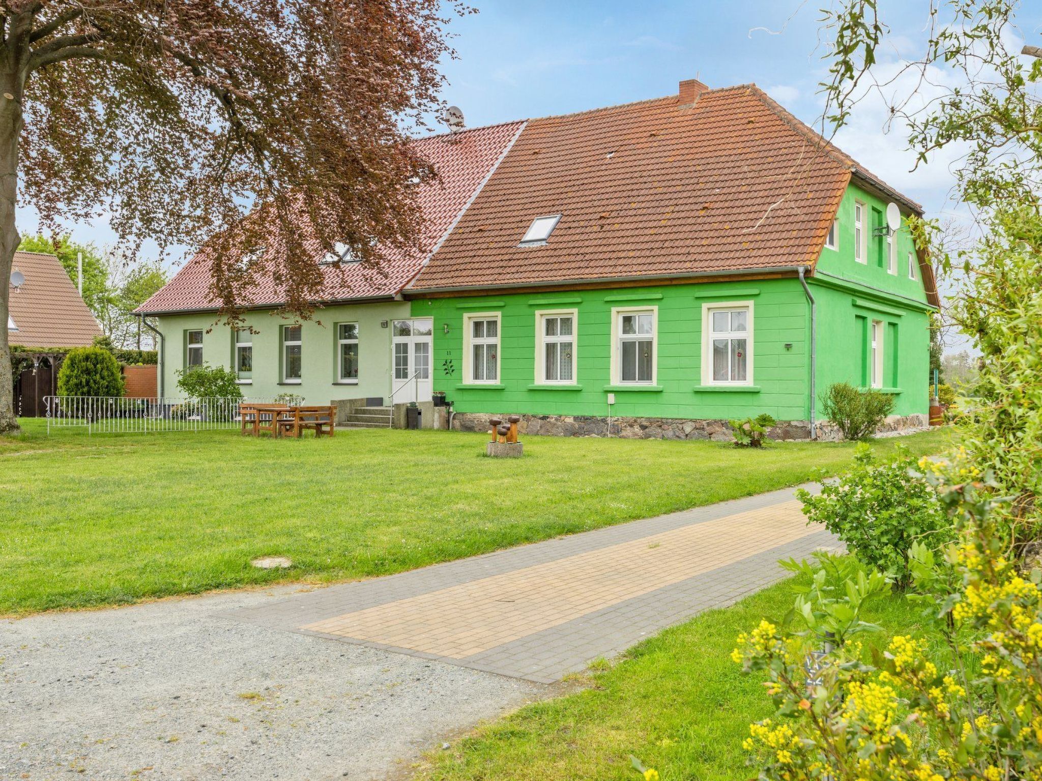 Une maison verte à deux étages, avec un toit recouvert de tuiles rouges, une allée pavée et un petit jardin parsemé de fleurs jaunes.