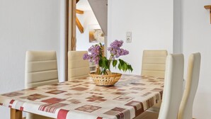 Un coin repas avec une table recouverte d’une nappe à motifs, quatre chaises de couleur beige et un panier rempli de fleurs violettes.