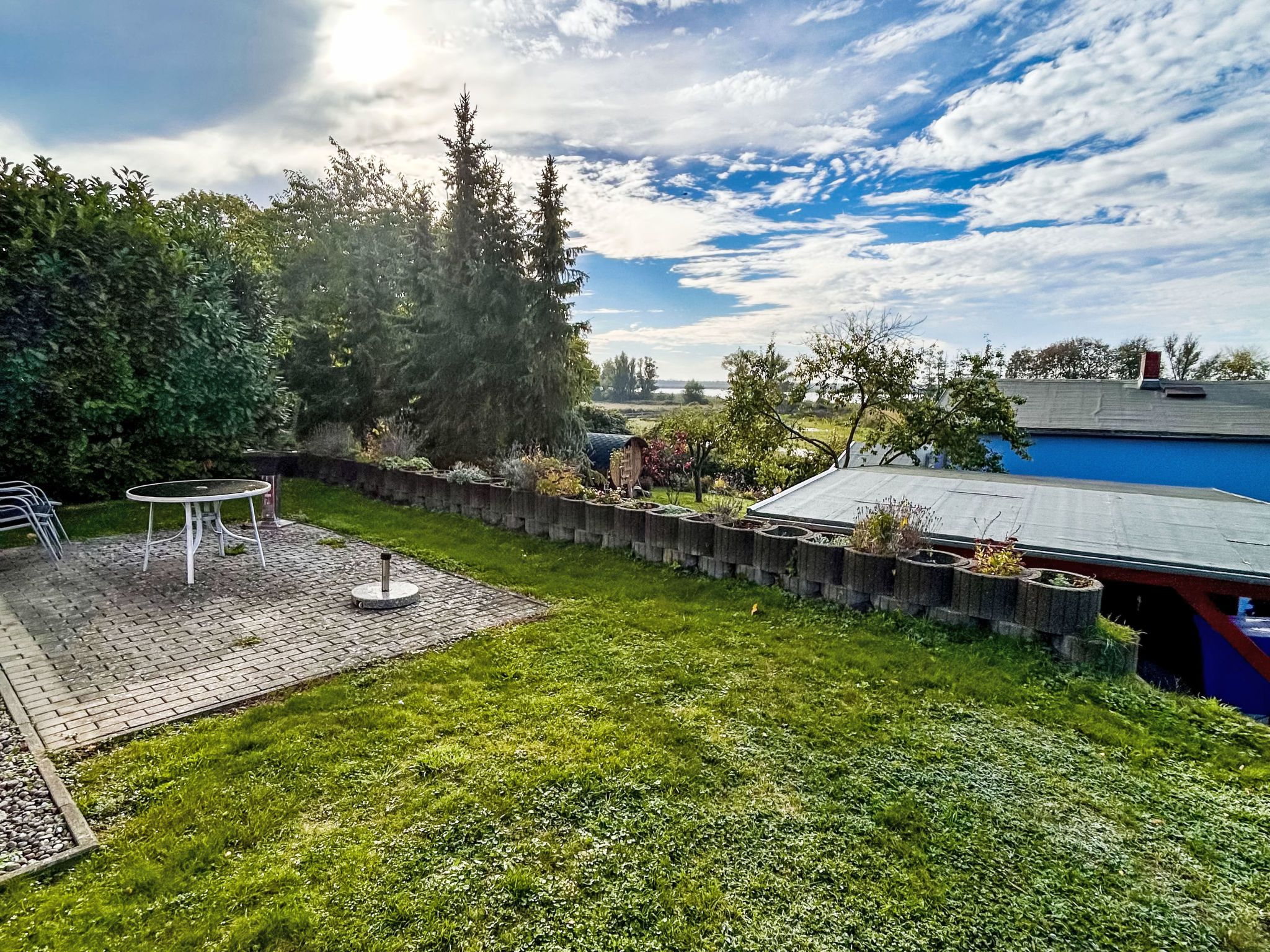 A backyard with a brick patio, a round table, and chairs. There is a wooden fence and various plants.