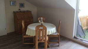 A dining area with a round table, two chairs, a wooden cabinet, and a window with a view of a deck.