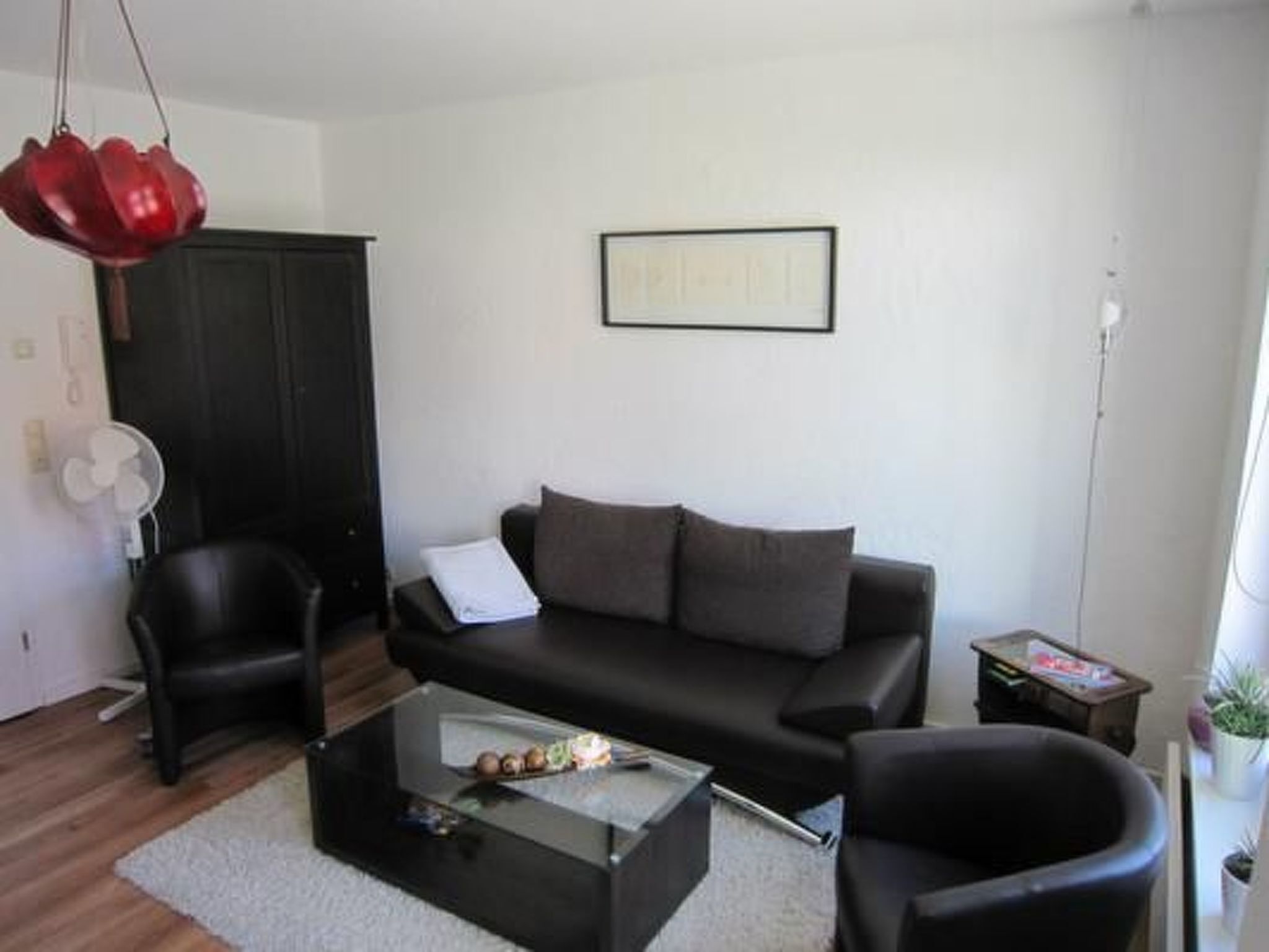 Ein modernes Wohnzimmer mit einer schwarzen Couch, zwei schwarzen Sesseln, einem Glastisch und einer roten Pendelleuchte.