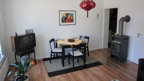 A dining area with a table and chairs, a hanging red light fixture, a television on a stand, a potted plant, and a wood-burning stove. - Charmante Wohnung in Malchow by Interhome (Göhren-Lebbin)