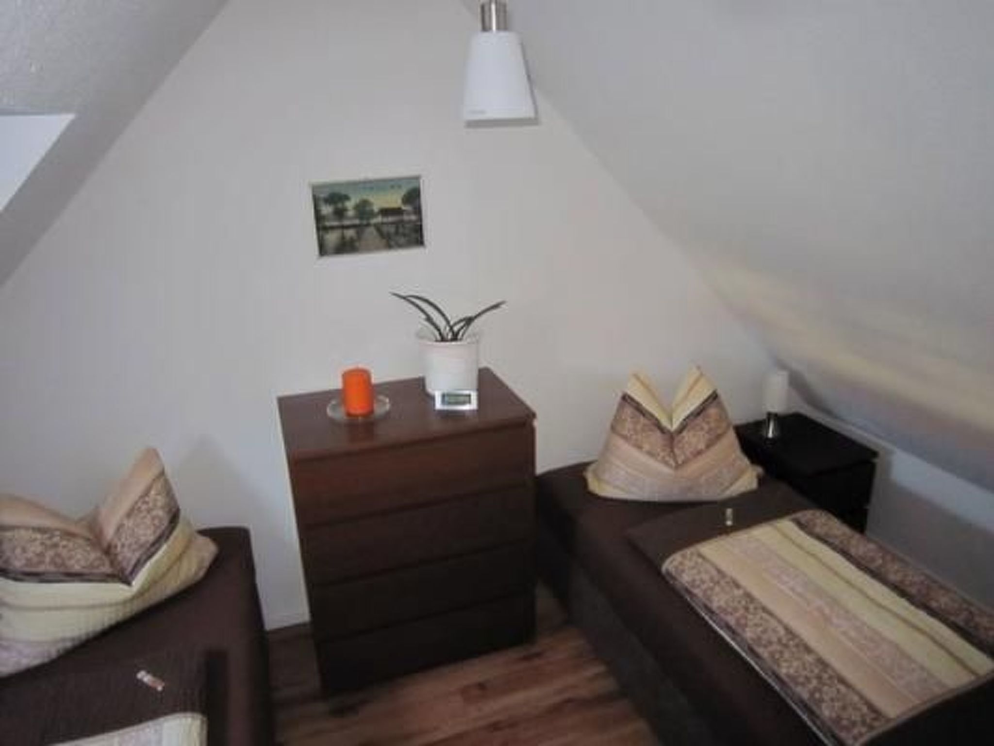 A small, cozy room with a sloped ceiling, a wooden floor, a small bedside table with a lamp, a framed picture on the wall, and two beds with pillows.