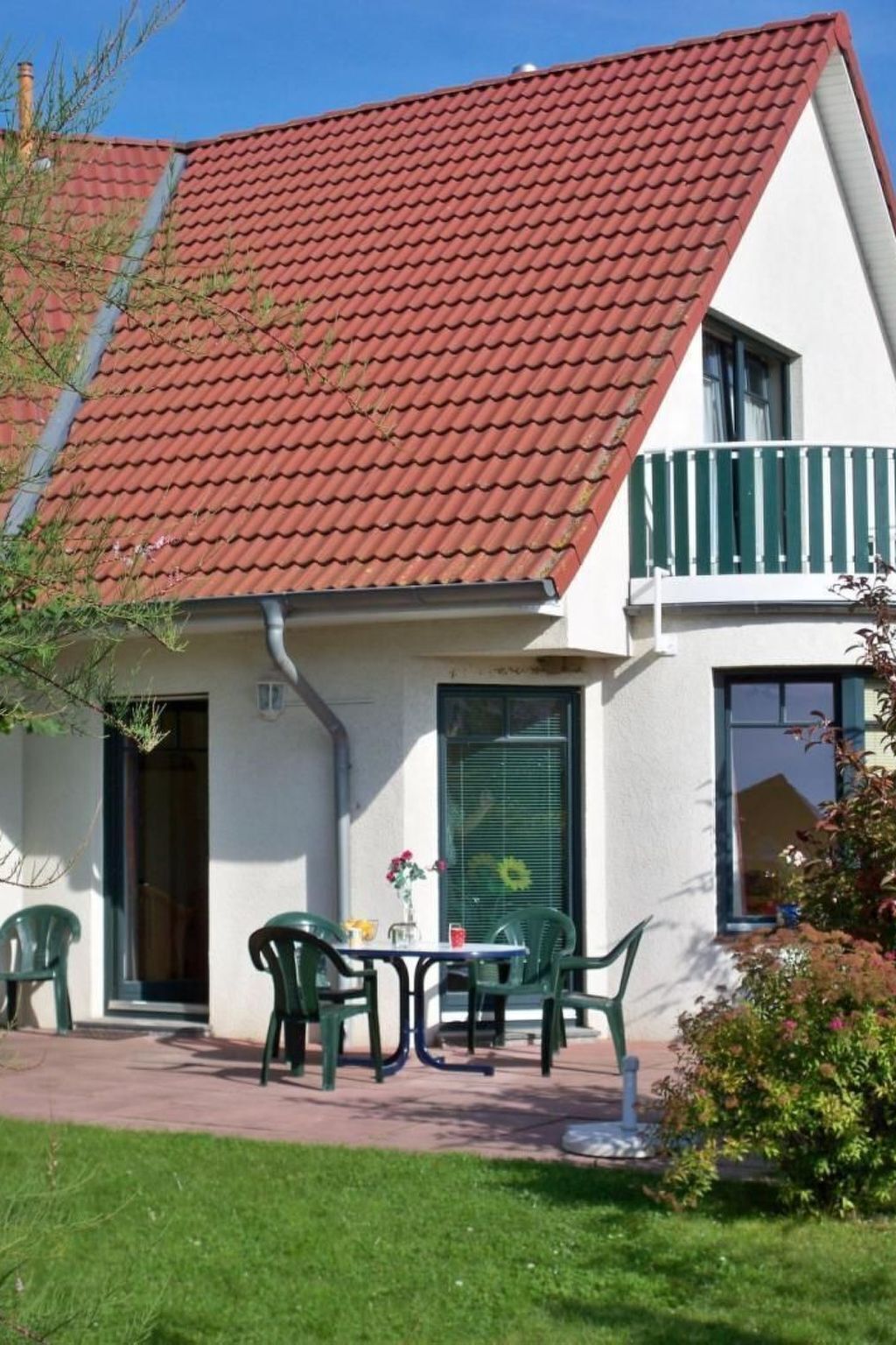 A two-story house with a red-tiled roof, a balcony with green railings, and a small patio area with outdoor furniture.
