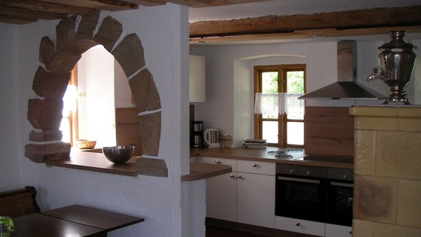 Una cocina con un arco de piedra, vigas de madera, un ventanal con cortinas de encaje y una estufa tradicional.