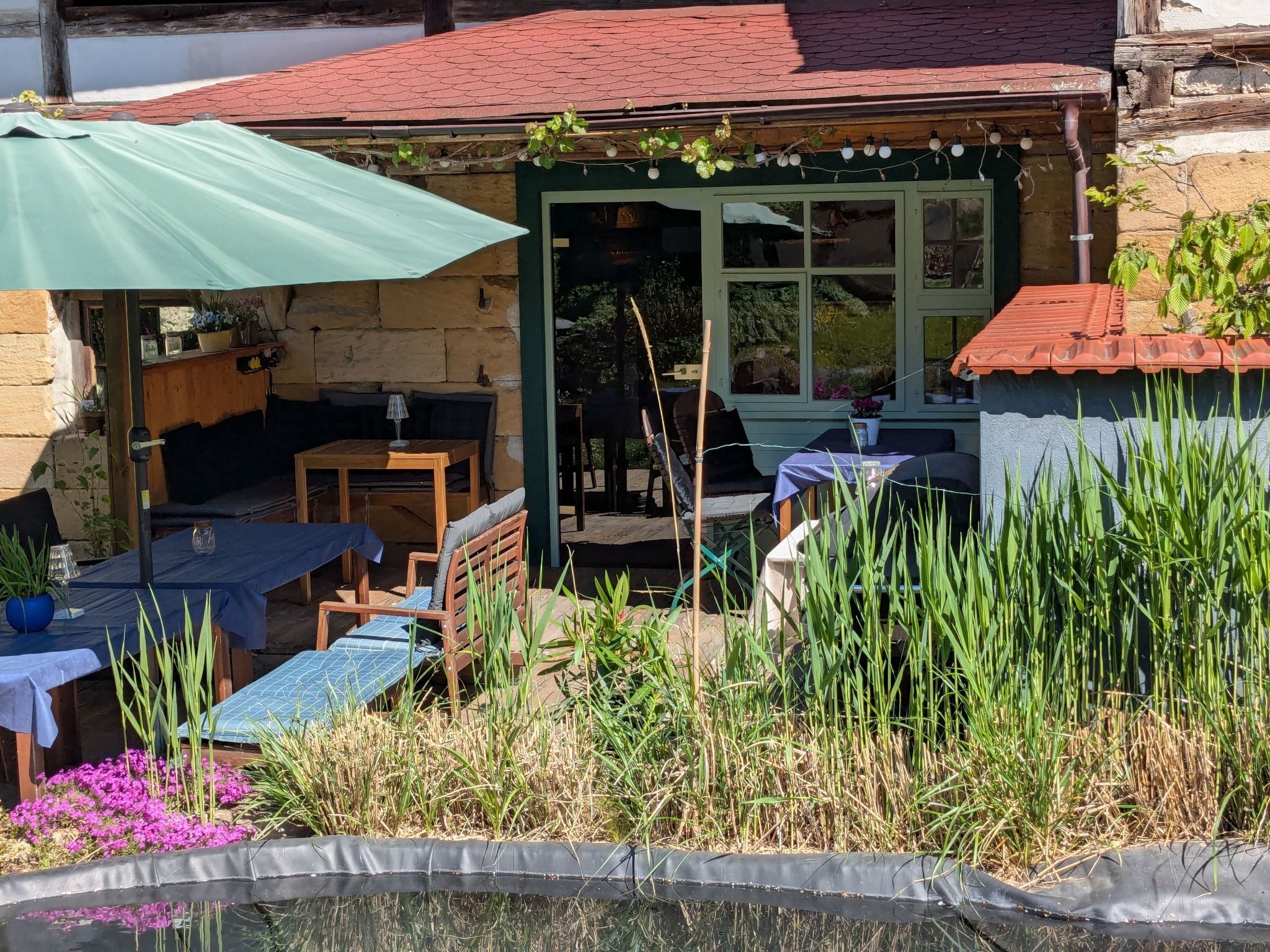 An outdoor seating area with tables and chairs, a small pond, and a stone building with a green door.
