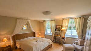 A bedroom with a large bed, bedside tables with lamps, a small chair, a bookshelf, and a window with curtains. - Apartment 45 qm by Interhome (Ahrenshoop)