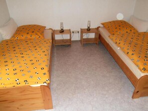 Il s’agit d’un petit logement composé d’une seule pièce, avec deux lits en bois, chacun recouvert d’une couverture jaune à motifs. On y trouve deux tables de chevet avec des lampes, et le sol est recouvert d’une moquette.
