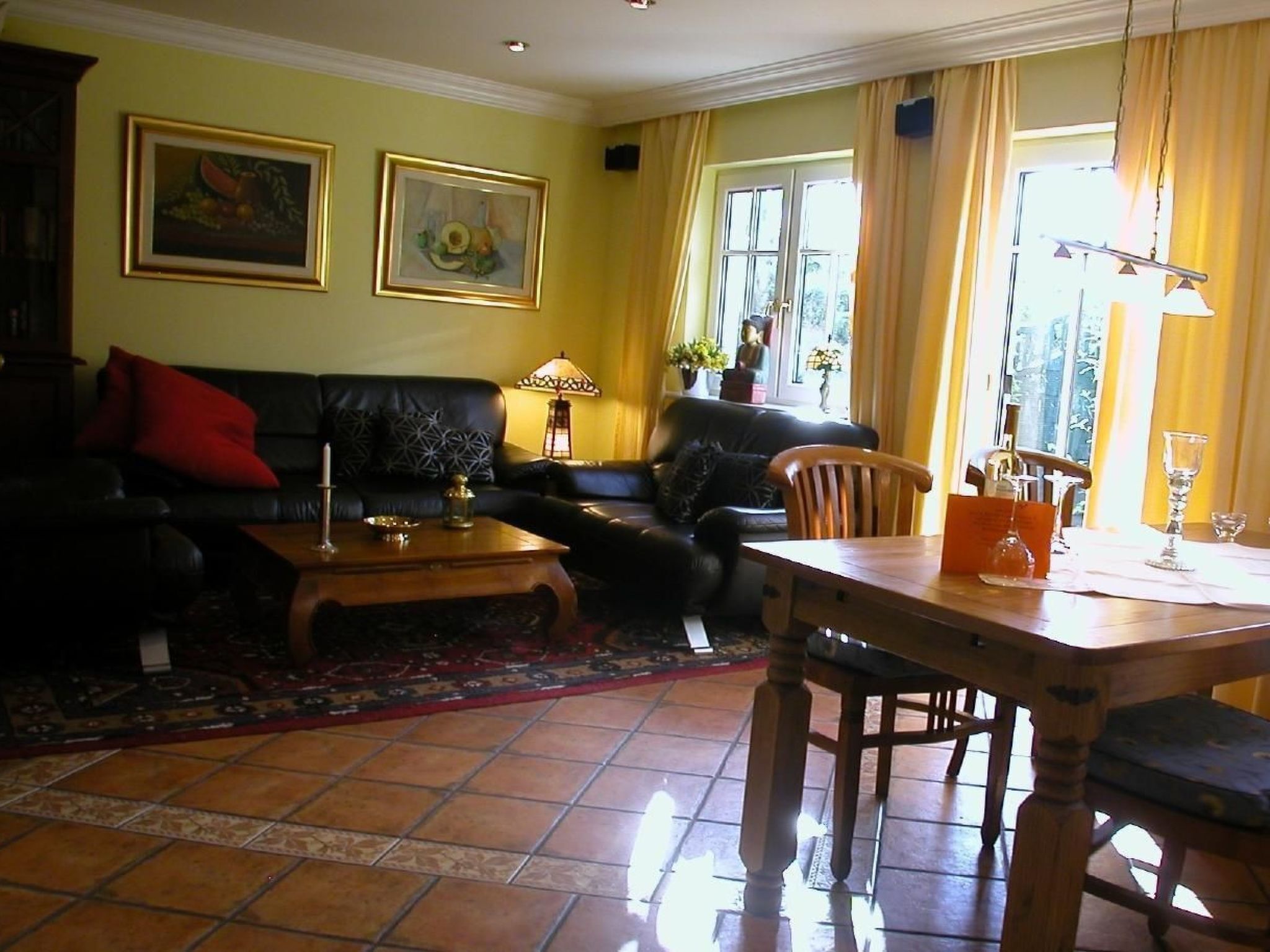 Ein Wohnzimmer mit einer Couch, einem Couchtisch und einem Esstisch mit Stühlen. An der Wand hängen zwei gerahmte Bilder, auf dem Couchtisch steht eine Lampe und es gibt ein Fenster mit Vorhängen.