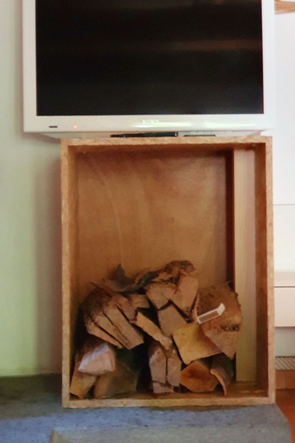 A television mounted on a wall with a wooden box below it containing firewood.