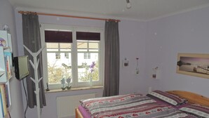 Una camera da letto con un letto, una finestra con tende, un termosifone, una TV su un mobile e un quadro appeso al muro.