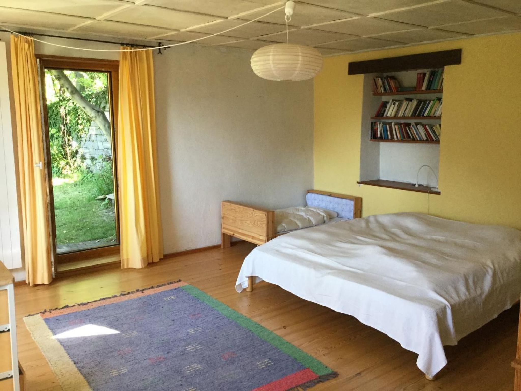 A bedroom with a bed, a bedside table, a bookshelf, and a window with curtains.