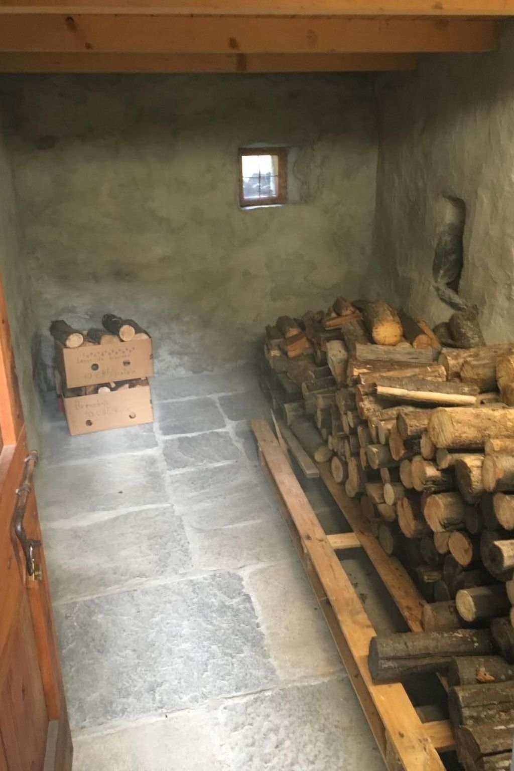 A narrow stone-walled room with a small window, wooden benches, and stacks of firewood.
