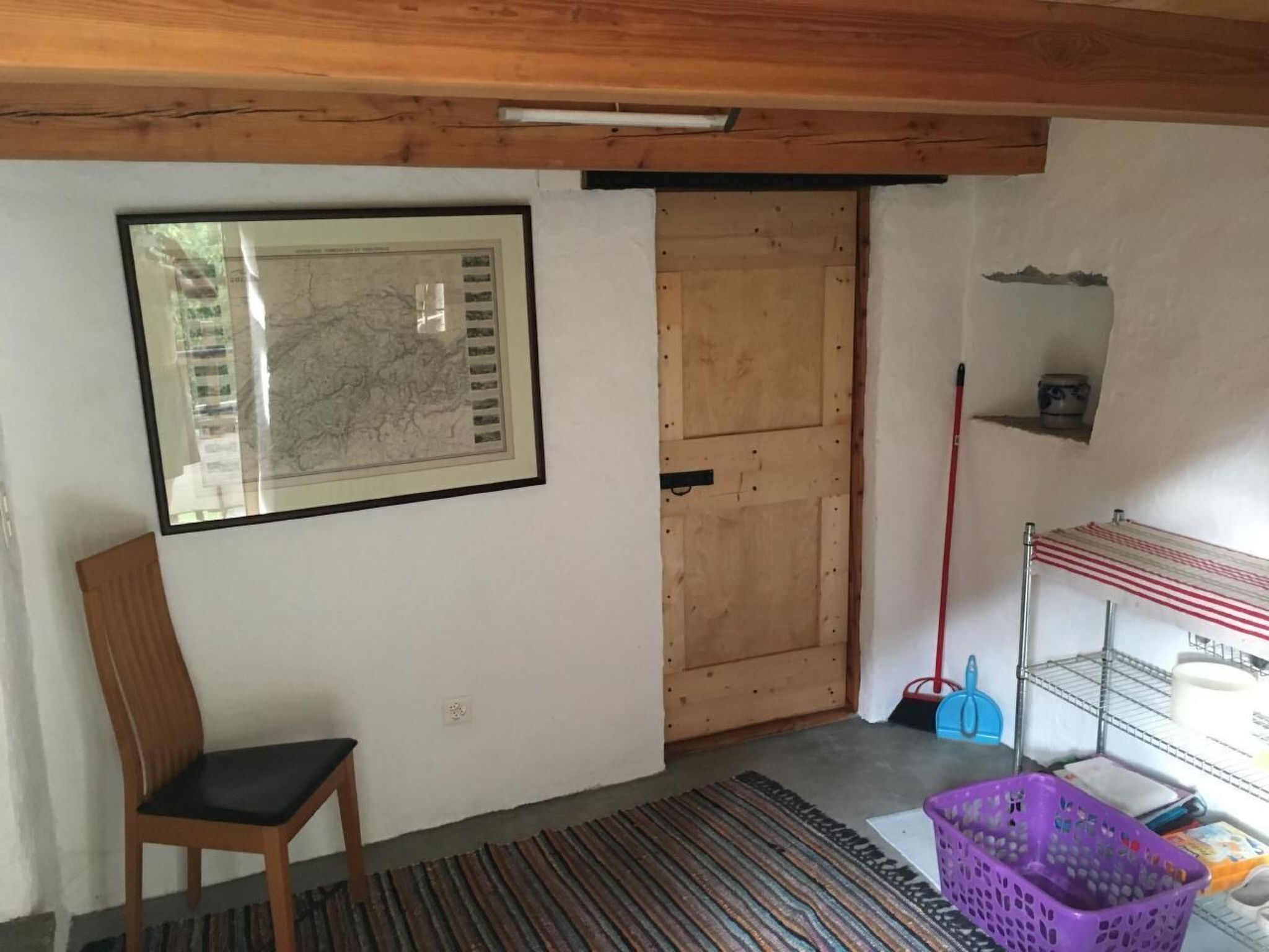 Ein Zimmer mit Holztür, einer gerahmten Karte an der Wand, einem Stuhl, einem violetten Wäschekorb und einem Metallregal mit Reinigungsmitteln.