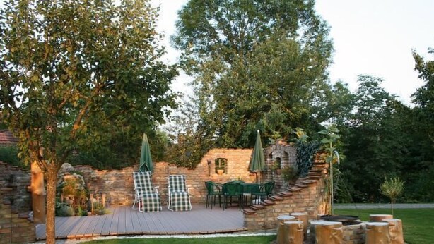 Een terras buiten met een stenen muur, een groen gazon en een zithoek met stoelen en tafels.