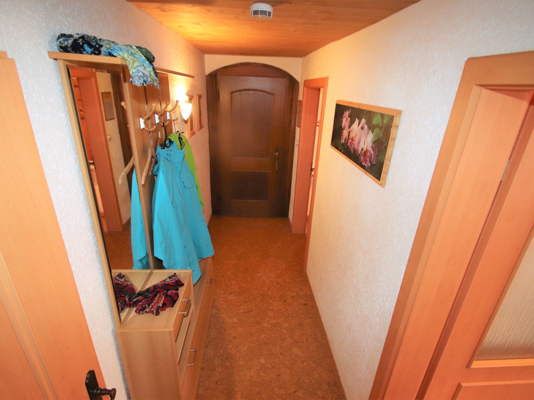 A narrow hallway with wooden doors, a coat rack with clothes, and a framed picture on the wall.