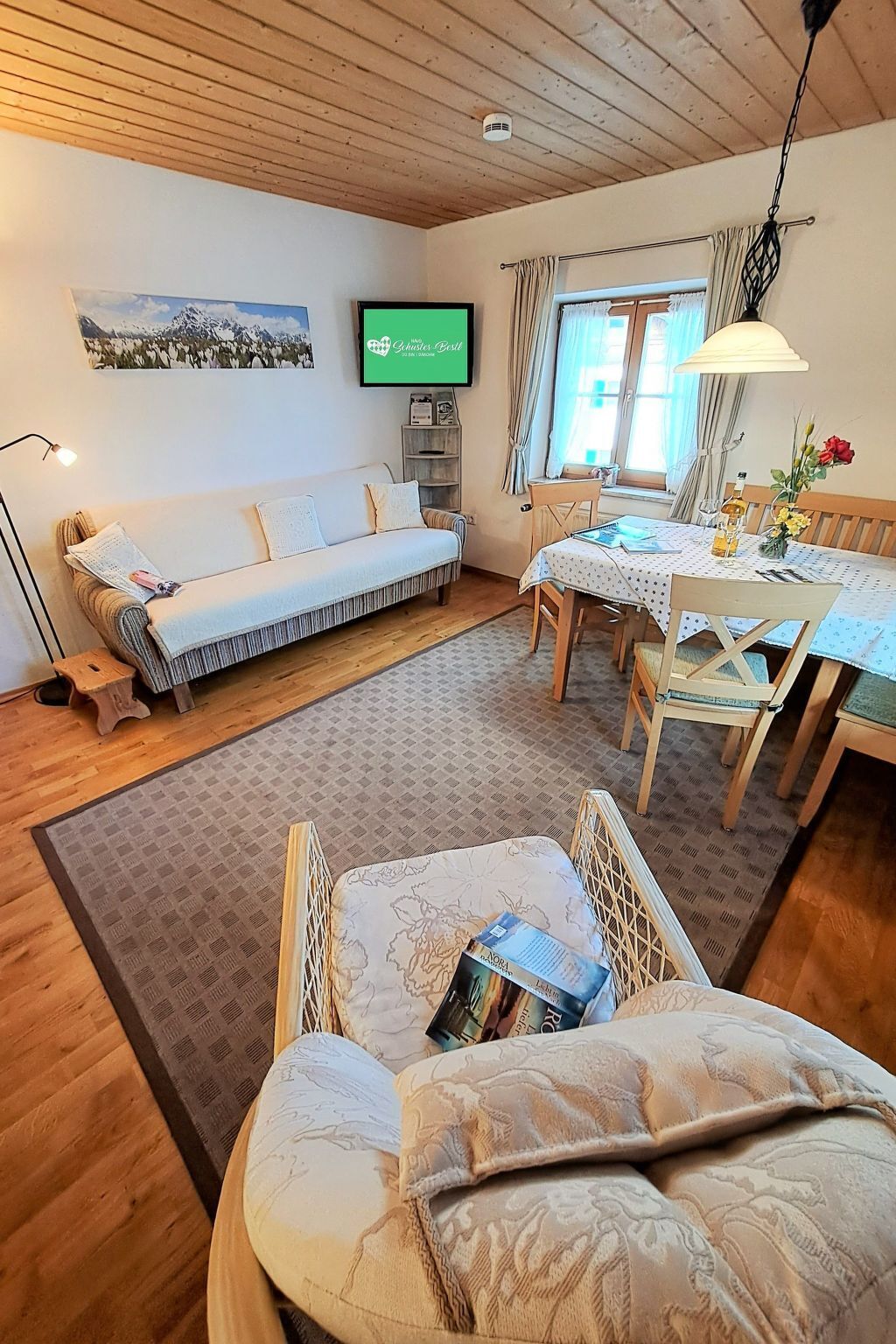 Un saló d'estar acollidor amb un sofà, una taula de menjador amb cadires, un televisor penjat a la paret i una finestra amb cortines.