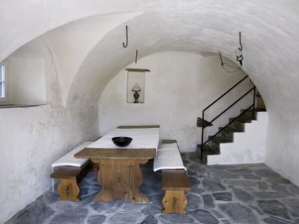 A stone-walled room with a wooden table and benches, a small window, and a staircase.