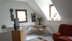 A small, well-lit room with a sloped ceiling, a desk with a computer, two chairs, a red sofa, and a small table with a plant.
