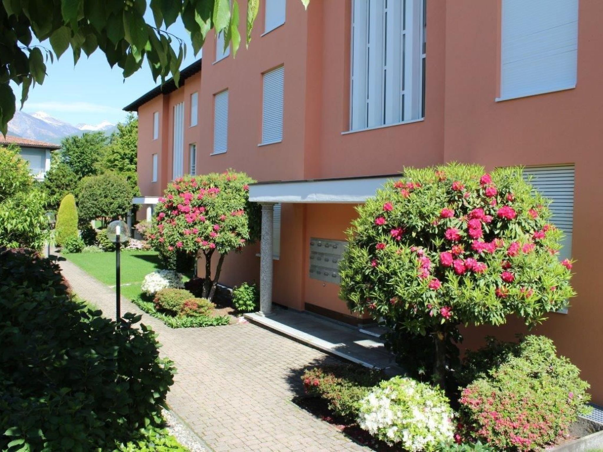A building with a paved pathway, surrounded by well-maintained hedges and flowering bushes.