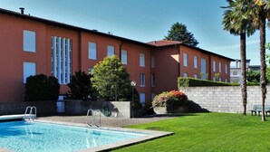 A swimming pool with a clear blue water, surrounded by a well-maintained lawn and a building with large windows and a red facade. - Residenza al Mulin by Interhome (Ascona)