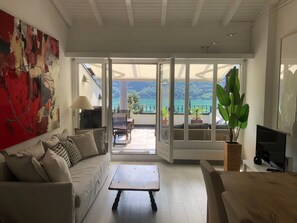 Ein modernes Wohnzimmer mit einer Couch, einem Couchtisch und einem Fernseher. Ein Balkon bietet Blick auf das Meer und die Berge.