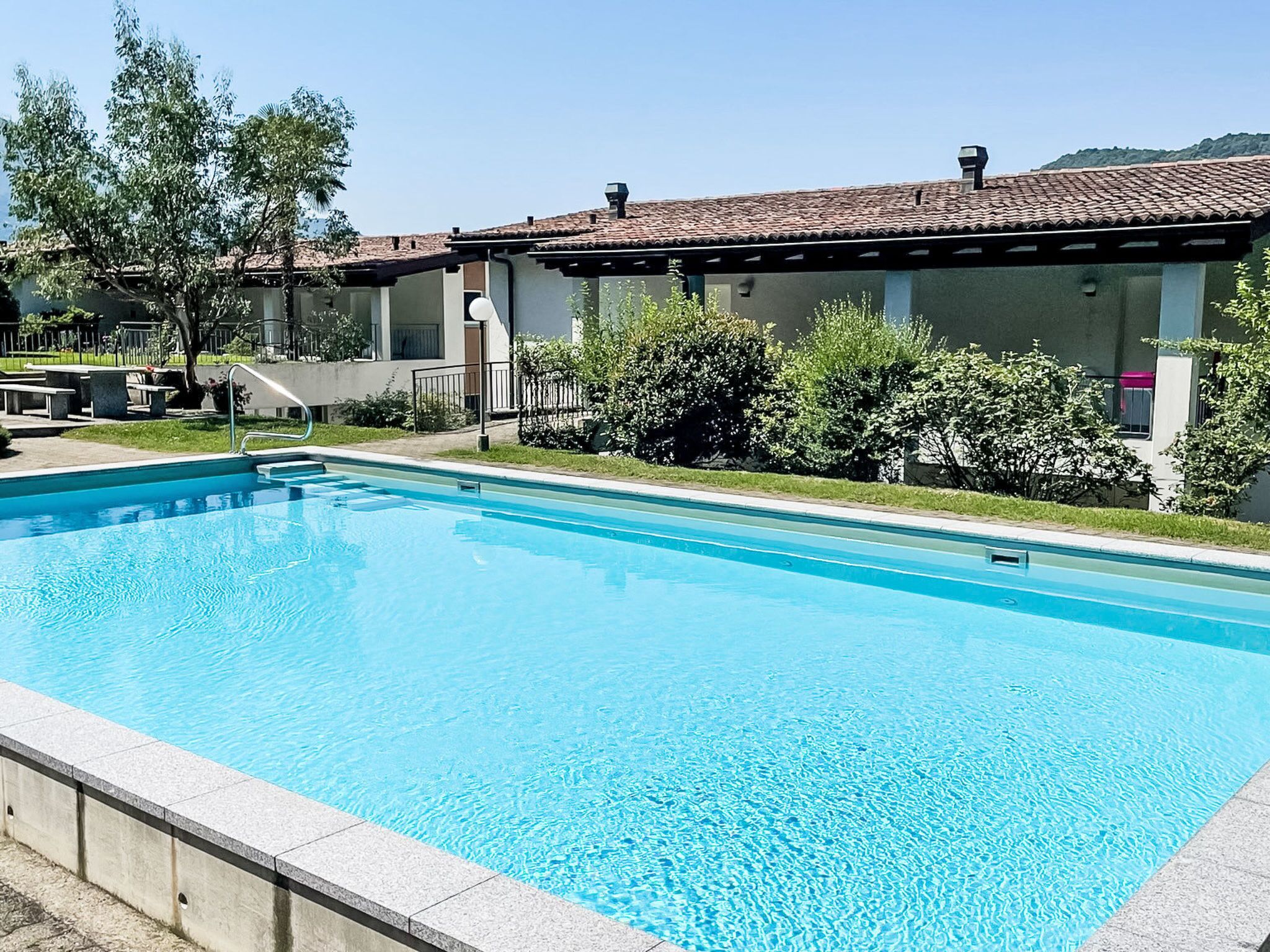 A swimming pool with a tiled deck, surrounded by greenery and a building with a tiled roof.